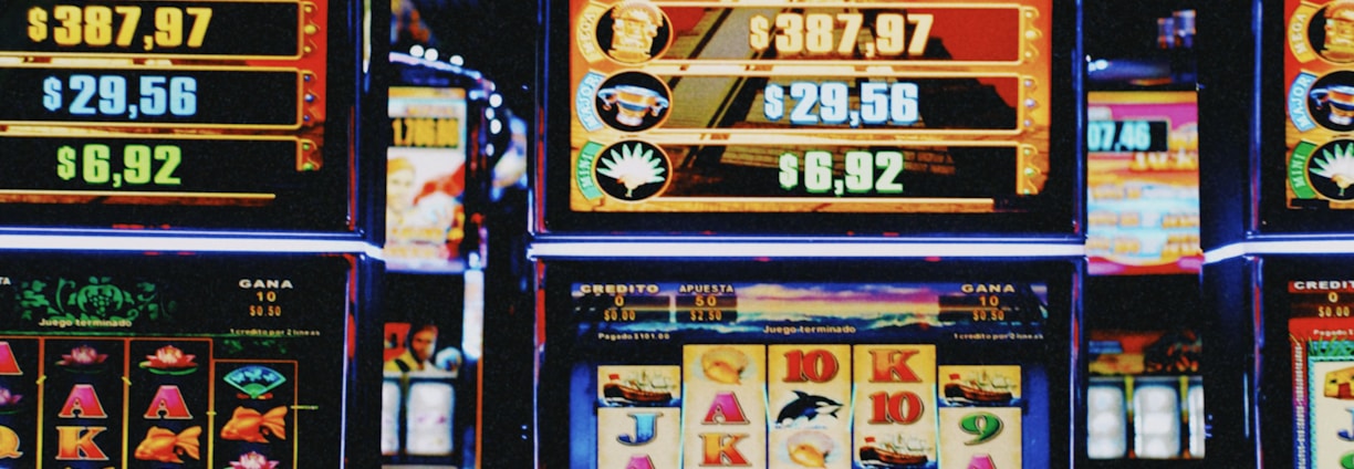 A row of colorful slot machines lit with bright neon lights, showcasing various graphical elements on the screens, indicating different games and jackpots. The atmosphere suggests a lively casino setting, bustling with excitement.